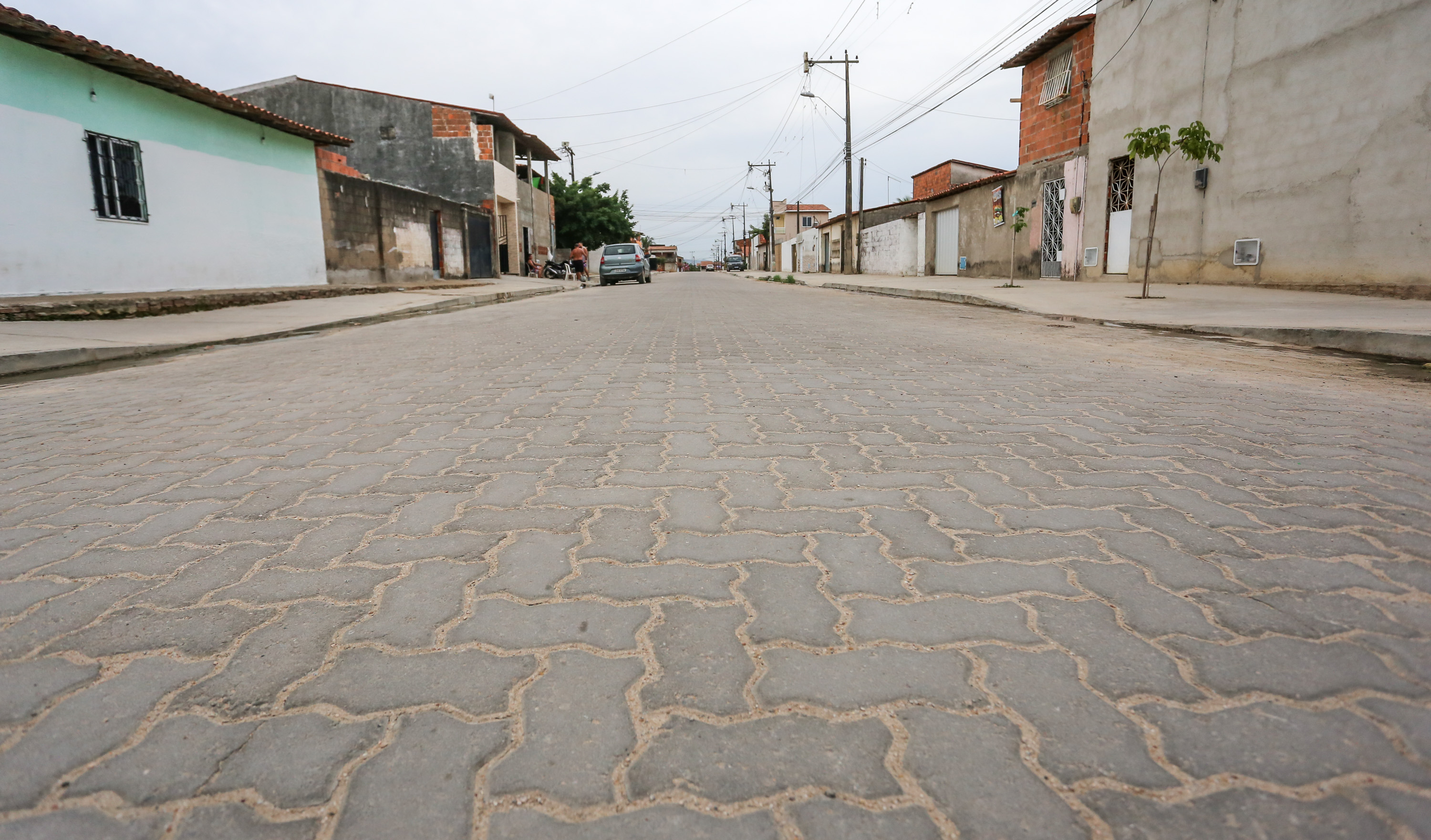 rua já concluída toda em piso intertravado e casas ao redor com um carro estacionado à esquerda e pessoas em pé ao lado